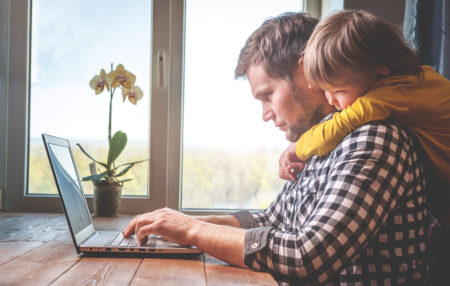 photo of a man using his computer, while his son hugs him